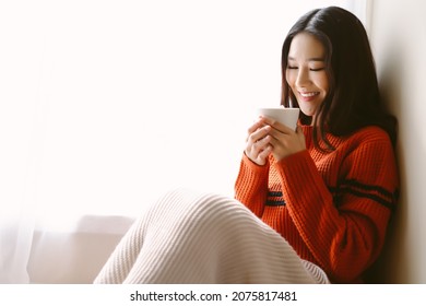 Portrait of beautiful Asian woman relaxing with cup of hot cocoa or chocolate with marshmallow while sitting by the window. Happy girl enjoying warm beverage in winter day.  Lazy day off concept. - Powered by Shutterstock