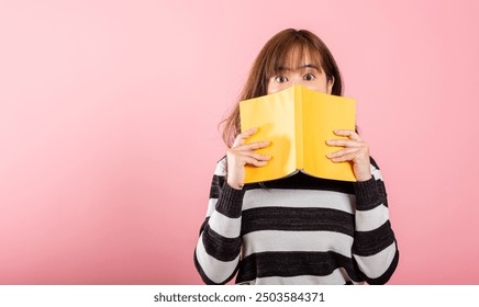 Portrait of beautiful Asian young woman teen smile covering her face with yellow book, female person hiding behind an open book show only eyes, studio shot isolated on pink background - Powered by Shutterstock