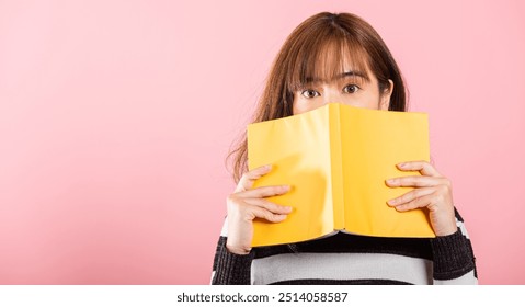 Portrait of beautiful Asian young woman teen smile covering her face with yellow book, female person hiding behind an open book show only eyes, studio shot isolated on pink background - Powered by Shutterstock