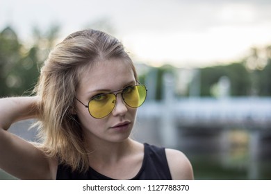 Portrait Of A Beautiful Attractive Cute Blond Girl In Yellow Aviators Glasses Straightening Hair And Looking At The Camera Outdoors In The Park