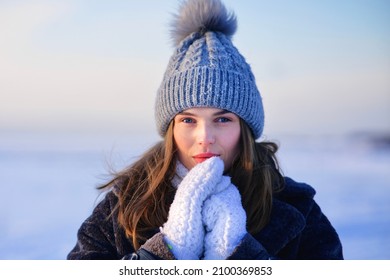 Portrait of beautiful attractive girl, young pretty woman standing, walking outdoors at cold winter frosty day on natural snow background in warm clothes knitted hat, coat and gloves looking at camera