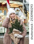 Portrait of beautiful attractive smiling slavic young girl in a knitted hat and mittens with fir tree branches enjoying holiday Christmas market, Winter, snowy day, festive city new year atmosphere