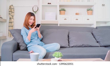 Portrait Of Beautiful Attractive Young Smiling Asian Woman Using Smartphone While Lying On The Sofa When Relax In Living Room At Home. Enjoying Time Lifestyle Women At Home Concept.