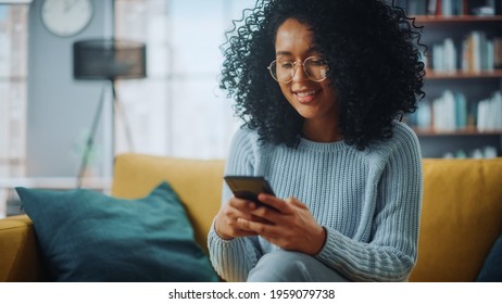 Portrait Of A Beautiful Authentic Latina Female In A Stylish Cozy Living Room Using Smartphone At Home. She's Browsing The Internet And Checking Videos On Social Networks And Having Fun.