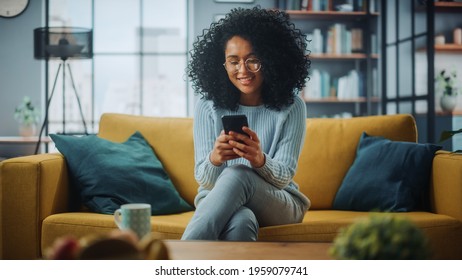 Portrait Of A Beautiful Authentic Latina Female In A Stylish Cozy Living Room Using Smartphone At Home. She's Browsing The Internet And Checking Videos On Social Networks And Having Fun.