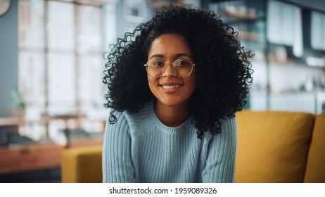 Portrait of a Beautiful Authentic Latina Female with Afro Hair Wearing Light Blue Jumper and Glasses. She Looks to the Camera and Smiling Charmingly. Successful Woman Resting in Bright Living Room. - Powered by Shutterstock