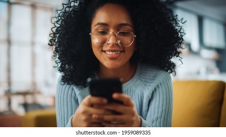 Portrait Of A Beautiful Authentic Latina Female With Afro Hair In Stylish Cozy Living Room Using Smartphone At Home. She's Browsing The Internet And Checking Videos On Social Networks And Having Fun.