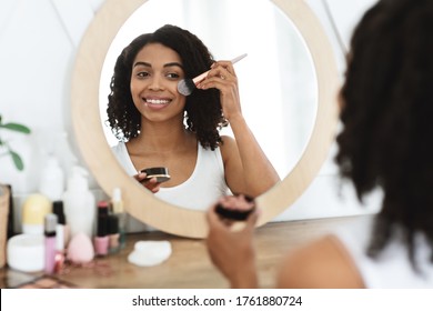 Portrait Of Beautiful Black Woman Applying Blush On Face, Using Brush Tool, Putting Makeup, Looking In Mirror At Home, Over Shoulder View, Selective Focus