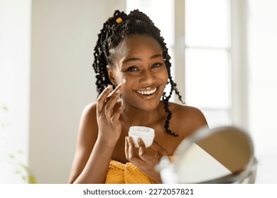 Portrait of beautiful black woman applying facial cream and smiling, lady moisturizing skin with lifting nourishing day creme after morning shower