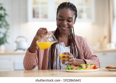 Portrait Of Beautiful Black Woman Eating Healthy Breakfast In Kitchen At Home. Happy African American Lady Sitting At Table, Enjoying Tasty Food And Pouring Orange Juice From Jug To Glass, Copy Space