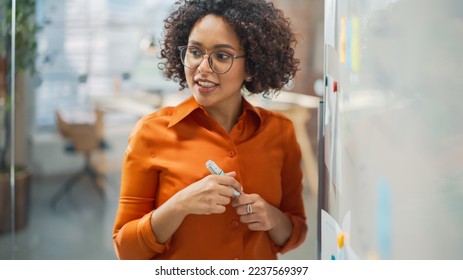 Portrait of a Beautiful Black Woman in Smart Casual Clothes Doing a Presentation in a Meeting Room for her Colleagues. Female Team Lead Explaining Data and Statistics Using a Whiteboard and a Marker - Powered by Shutterstock