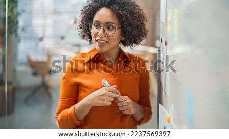 Portrait of a Beautiful Black Woman in Smart Casual Clothes Doing a Presentation in a Meeting Room for her Colleagues. Female Team Lead Explaining Data and Statistics Using a Whiteboard and a Marker