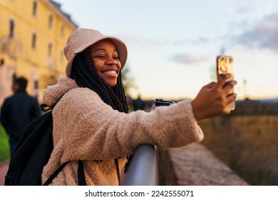 Portrait of a beautiful black woman smiling excitedly at the camera, she's wearing a bucket hat and a backpack, she's drinking coffee and taking pictures with her phone - Powered by Shutterstock