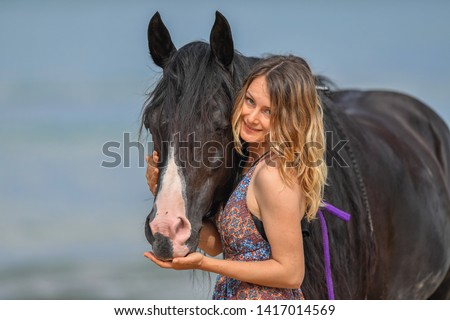 Portrait of a beautiful blond girl hugging with her black horse on the sea beach