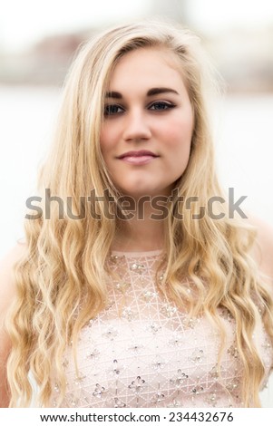Portrait of a beautiful blond teenage girl with wavy long hair, blue eyes, pink lips and pink dress standing isolated alone outdoors.