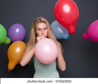 Portrait of a beautiful blond woman blowing up balloon on gray background