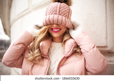 Portrait of beautiful blond woman dressed in pink warm clothes, padded jacket, white sweater, pants and knitted hat with fur pumpon walking in the street of the city. Urban style concept