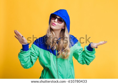 portrait of beautiful blond woman in sunglasses and blue green hooded jacket on yellow background. hipster summer.