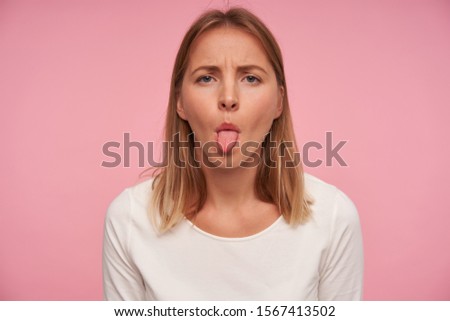Portrait of beautiful blonde female with casual hairstyle frowning eyebrowns and showing her tongue while posing over pink background, wearing white blouse