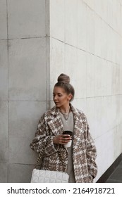 Portrait Of Beautiful Blonde Girl Dressed In Oversize Beige Coat With Print, Knitted Dress, White Bag In Hands, Hair Gathered In Bun, Stylish Trendy Fashion Outlook, Lifestyle Model