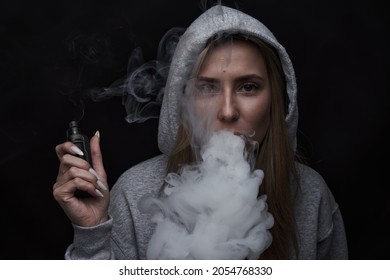 Portrait Of Beautiful Blonde Girl In Gray Hoodie Smokes Vape Isolated On Black Studio Background, Cloud Of Steam Smoke, Mini Hookah