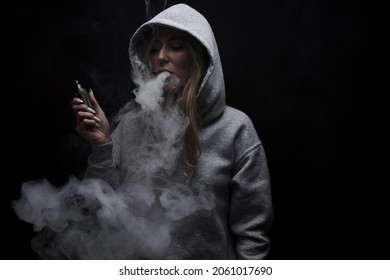 Portrait Of Beautiful Blonde Girl In Gray Hoodie Smokes Vape Isolated On Black Studio Background, Cloud Of Steam Smoke, Mini Hookah