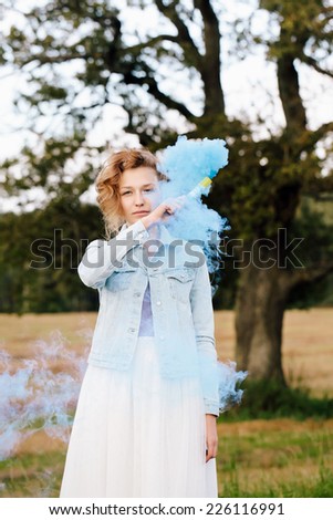 Portrait of beautiful blonde girl keeping blue smoke popper in her hand near her face. Nature background. Outdoors