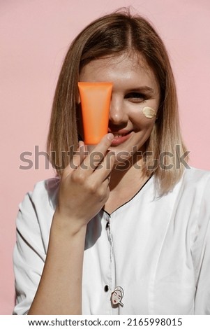 portrait of beautiful blonde girl with plump lips, model hides face behind an orange jar of cream, holds tube near eye and smiles, cream on cheek, sun glare and shadows on face, advertising, close up