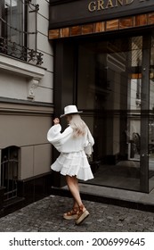 Portrait Of A Beautiful Blonde Girl In A White Dress And Hat, Light Stylish Summer Fashion Look, Ancient Building On The Background, Full Length Lifestyle Model Stands Backwards And Whirling