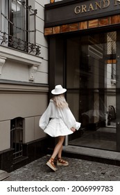 Portrait Of A Beautiful Blonde Girl In A White Dress And Hat, Light Stylish Summer Fashion Look, Ancient Building On The Background, Full Length Lifestyle Model Stands Backwards And Whirling