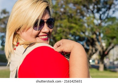 portrait beautiful blonde plus size latin argentinian young woman with sunglasses smiling outdoors looking at the camera, on a sunny day in the park with trees in the background, copy space.