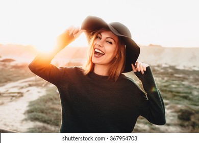 Portrait of beautiful blonde young woman in glasses and holding hat and winking on the beach with sunset light. 