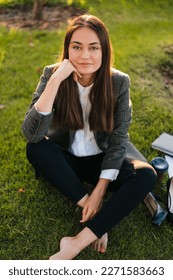 Portrait of a beautiful brunette business woman sitting on the grass barefoot and smiling while looking at camera enjoying the work break in the park. eople
