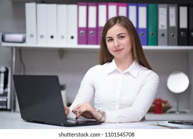 Portrait of beautiful brunette girl working in office behind laptop on desk. She sits right in front of the camera smiling and looks happy