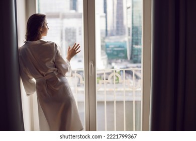 portrait of a beautiful brunette woman in a bathrobe stands at a large window in an apartment opposite a skyscraper in the morning - Shutterstock ID 2221799367