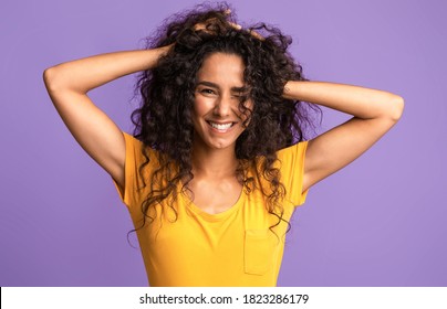 Portrait of beautiful brunette woman touching her natural curly hair and smiling, standing isolated on purple background, looking and winking at camera, closeup shot of cheerful young lady, copy space