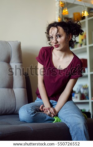 Portrait of beautiful brunette young woman with curly funny hairstyle sitting on the couch, wearing blue jeans and wine red t-shirt, has a perfect makeup, mirror on the background