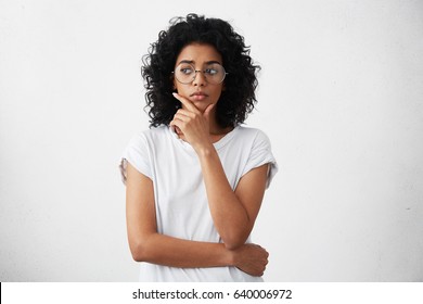 Portrait of beautiful casually dressed young woman in round glasses having doubtful expression, looking away in indecisiveness, holding her chin, trying to find best solution. Body language