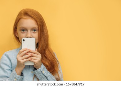 Portrait Of Beautiful Caucasian Red Haired Kid Girl With In Hands, Surprised Girl Hold Mobile Phone Near Her Face And Look At Camera. Isolated Yellow Background