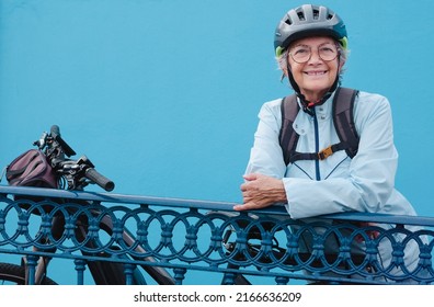 Portrait Of Beautiful Caucasian Senior Cyclist Woman Wearing Bike Helmet And Backpack Standing Close To Her Bicycle Looking Away Smiling. Blue Background, Copy Space