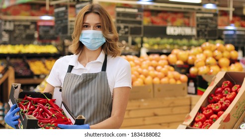 Portrait Of Beautiful Caucasian Woman In Face Mask And Apron Standing In Food Store With Red Hot Chili Papers Indoors. Pretty Female Seller Holding Box With Food In Supermarket. Retail Concept