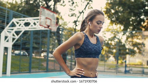 Portrait Of Beautiful Caucasian Young Slim Woman Smiling Cheerfully To Camera While Standing At Sport Court In Summer. Happy Charming Girl In Sporty Outfit Looking Straight. Female Model In Airpods.