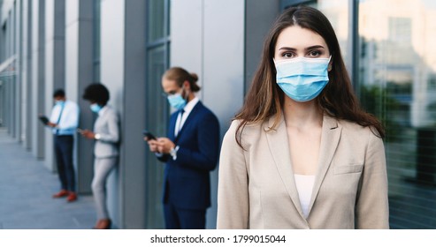 Portrait Of Beautiful Caucasian Young Woman In Medical Mask And Business Style Standing Outdoor And Looking At Camera. Line Of Mixes-races People With Gadgets On Background. Pandemic Concept.