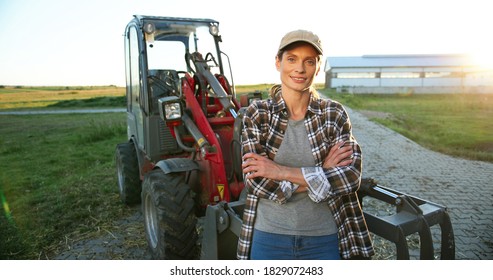 Portrait of beautiful Caucasian young woman in cap standing outdoors a big tractor machine and smiling cheerfully to camera. Pretty happy female farmer worker in field at farm. Agriculture.