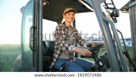 Portrait of beautiful Caucasian young woman in cap sitting in big tractor machine and smiling cheerfully to camera. Pretty happy female farmer worker in field at farm. Agricultural work.