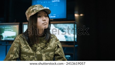 Portrait of beautiful Caucasian young woman in camouflage uniform and hat sitting at desk and working at computer, then turning face to camera and looking straight Female in military monitoring center