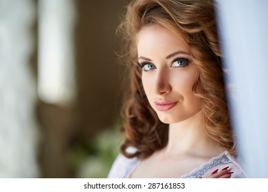 Portrait of a beautiful charming girl with blond curly hair, blue eyes, big full lips and curvy Boobs in white underwear and a transparent negligee with passionate look from the curtains at the window