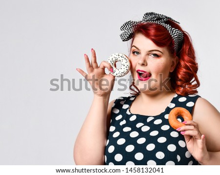 Portrait of beautiful cheerful fat plus size woman pin-up wearing a polka-dot dress isolated over light background, eating a donut. Diet, dieting concept