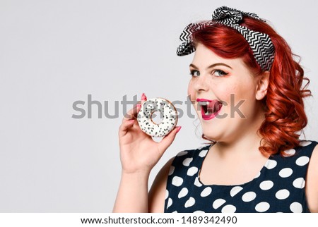 Portrait of beautiful cheerful fat plus size woman pin-up wearing a polka-dot dress isolated over light background, eating a donut.
