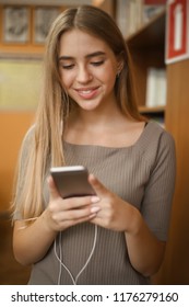 Portrait Of A Beautiful Cheerful Smiling Young Student Schoolgirl Lady With Long Hair Standing In Empty Classroom Listening Music With Earphones Using Mobile Phone Chatting.